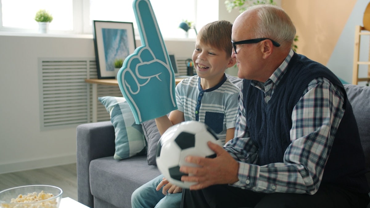 Cheerful family elderly grandfather and grandson watching soccer on TV cheering having fun together. Sports, entertainment and leisure time concept.